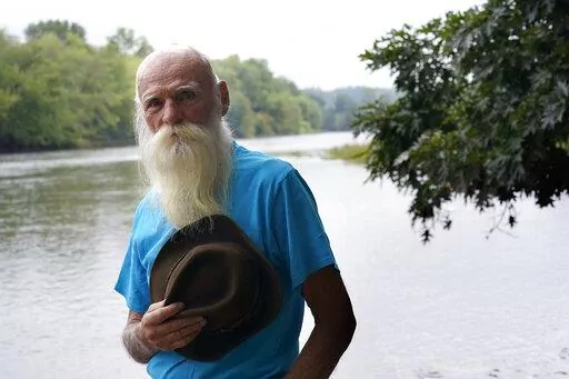FILE — David Lidstone, 81, stands for a photograph near the Merrimack River, Tuesday, Aug. 10, 2021, in Boscawen, N.H. Lidstone, a former hermit in New Hampshire, known to locals as "River Dave," whose cabin in the woods burned down after nearly three decades on the property that he was ordered to leave, and who received more than $200,000 in donations, has been charged with trespassing there once again. Lidstone still disputes that he is on the property, and was arrested on a trespassing char