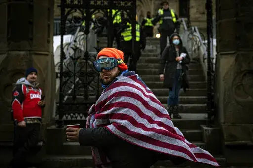 A man wearing an American flag walks by police guarding the Canadian parliament building from demonstrators protesting the country's COVID-19 restrictions, Wednesday, Feb. 16, 2022, in Ottawa, Ontario. The protests in Canada that have blocked border crossings with the U.S. and gnarled trade have been promoted, cheered and funded by American anti-vaccine groups, right-wing activists and conservative elected officials.  (AP Photo/Robert Bumsted)